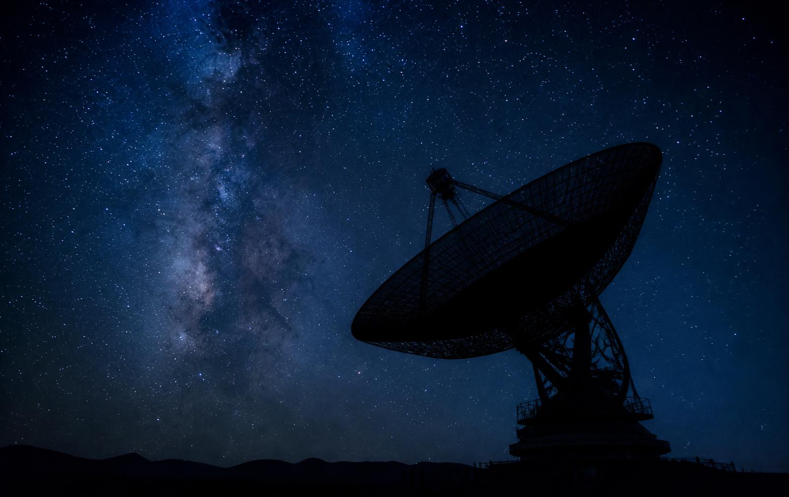 A radio telescope silhouetted against a star field and the Milky Way
